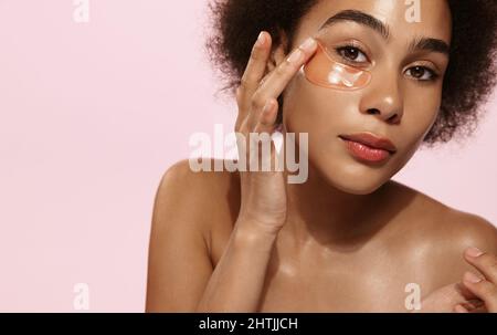 Primo piano di african american ragazza con naturale senza trucco aspetto e sorriso bianco, togliere il silicone sotto gli occhi cerotti e sorridere felice, utilizzando la cura della pelle Foto Stock