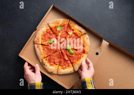 Deliziosa pizza Pepperoni in cartone. Mani maschili che raccolgono la fetta di pizza piccante Pepperoni, sfondo ardesia nero. Vista dall'alto. Consegna cibo Concep Foto Stock