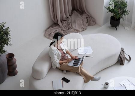Giovane donna d'affari asiatica con lapto pon le ginocchia concentrandosi sulla rete mentre si siede sul comodo divano bianco a casa Foto Stock