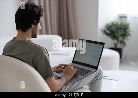 Giovane uomo contemporaneo con un computer portatile in ginocchio che guarda sullo schermo mentre si siede sul divano in salotto Foto Stock