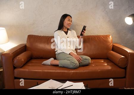 Giovane donna asiatica sorridente che comunica in videochat mentre si siede sul divano e tiene lo smartphone davanti a se stessa Foto Stock