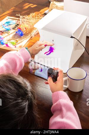 Dall'alto di raccolto anonimo artigiano femminile scattando foto di orecchini fatti a mano per i social media blog, mentre si utilizza lo smartphone e seduti in officina Foto Stock