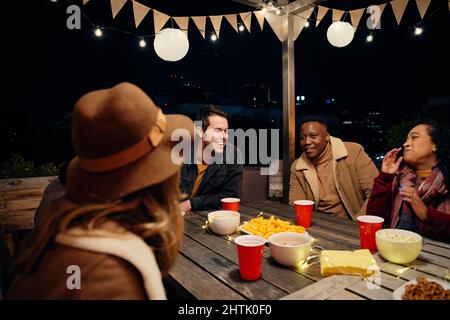 Gruppo multiculturale di giovani amici adulti che ridono mentre intorno ad un tavolo in una festa alla moda sul tetto. Mangiare e bere, avere un buon tempo Foto Stock