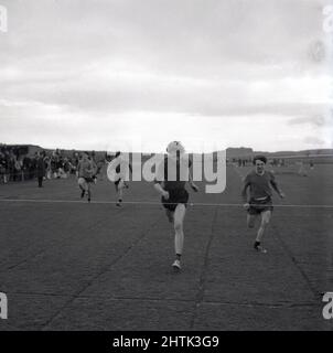 1960s, storico, atletica scolastica, all'aperto su un campo d'erba in una gara di corsa, due giovani maschi che si sprinting per il nastro di finitura, Fife, Scozia, Regno Unito. Foto Stock