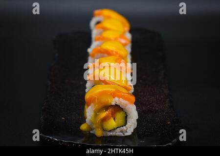 Combinazione tropicale di sushi tradizionale giapponese Uramaki con salmone selvatico, frutta fresca di mango e salsa di mango dolce. Sfondo nero. Foto Stock