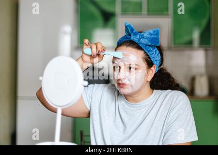 Giovane donna caucasica che mette maschera idratante sulla pelle del viso con spazzola di gomma che ha arco blu sulla testa guardando specchio. Parlare di cura di te e. Foto Stock