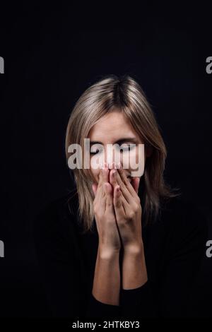 Donna spaventata con le mani che nascondono naso e bocca in silenzio vestita in blusa nera su sfondo nero. Vittima di abuso fisico e psicologico Foto Stock