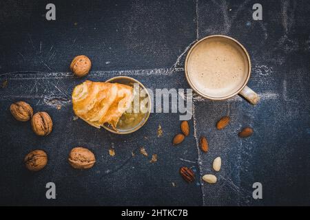 Un pezzo di delizioso croissant francese immerso nella marmellata, una tazza di caffè e alcune noci, scena della colazione, sfondo nero, vista dall'alto Foto Stock