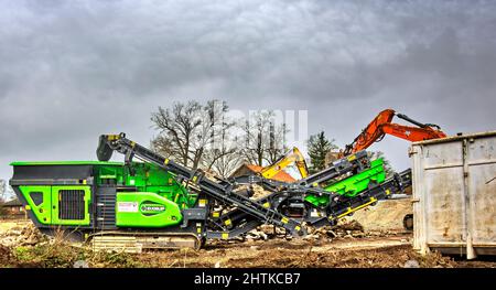 Gifhorn, Germania, 25 febbraio 2022: Trituratore con nastri trasportatori per materie prime minerali di fronte a un cumulo di materiale da demolizione da a contro Foto Stock