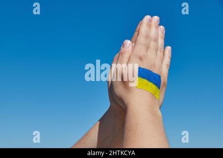 Le mani delle donne con il simbolo della bandiera ucraina pregano contro il cielo blu. Fermare la guerra Foto Stock