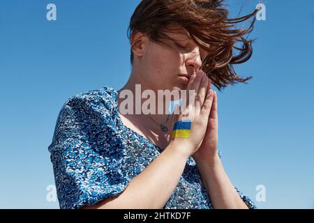 La donna Ucraina con un simbolo di bandiera in mano prega contro il cielo blu. Fermare la guerra Foto Stock