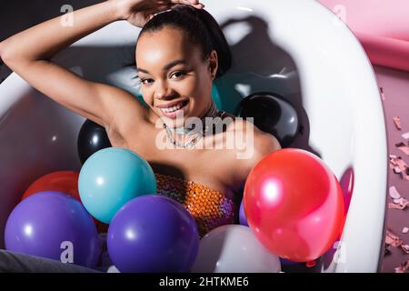 vista ad alto angolo della donna afro-americana soddisfatta che giace nella vasca da bagno con palloncini colorati Foto Stock