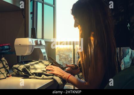bella giovane donna caucasica nella sua officina taglio tessuti seduti accanto alla finestra e la luce del sole delimita il suo viso, con una macchina da cucire sul Foto Stock