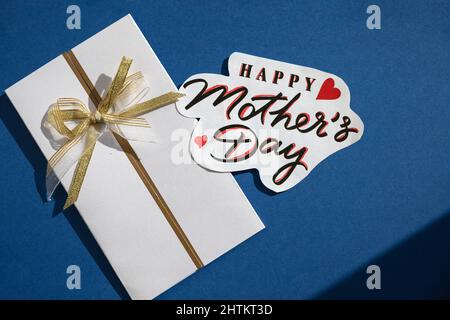 testo del giorno della mamma felice e busta bianca su sfondo blu, vista dall'alto. Elegante biglietto d'auguri, piatto laico. Mamme giorno. Primavera immagine Foto Stock