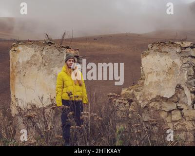 Una donna si erge vicino al muro di casa rovinata in erba secca alta sullo sfondo delle colline autunnali con mucche al pascolo e foschia nebbia Foto Stock