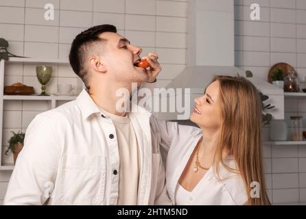 Bella donna che alimenta marito o ragazzo mentre cucinano insieme nella moderna cucina scandinava. Cena romantica, concetto di trascorrere del tempo insieme. Foto di alta qualità Foto Stock