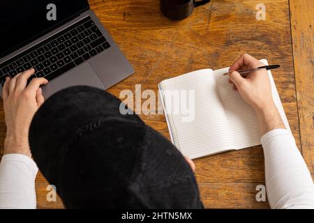 Vista dall'alto dell'uomo che scrive nel blocco note e utilizza il computer portatile alla scrivania Foto Stock