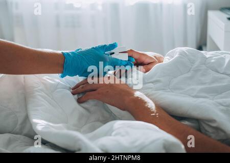 L'uomo si trova a letto, un medico in guanti medici si presenta a lui e mette un pulsossimetro sul suo dito per misurare la saturazione di ossigeno. Trattamento dell'asma Foto Stock