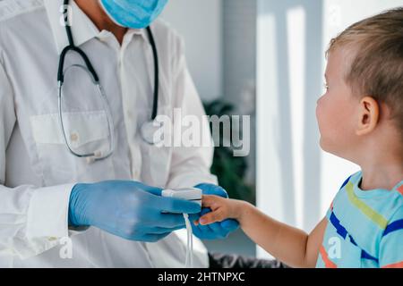 Un medico che indossa guanti medici e una maschera mette un pulsossimetro sul dito del bambino per misurare la saturazione di ossigeno. Il ragazzino controlla l'ossigeno Foto Stock