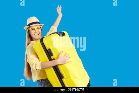 Ritratto di giovane donna felice in cappello che porta la sua valigia gialla su sfondo blu Foto Stock