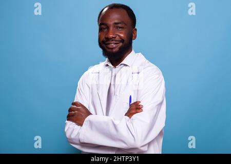 Ritratto di sorridente medico afroamericano in posa con le braccia incrociate in studio. Medico professionista residente in cerca di confindent in piedi in bianco camice da laboratorio. Professionista sanitario. Foto Stock
