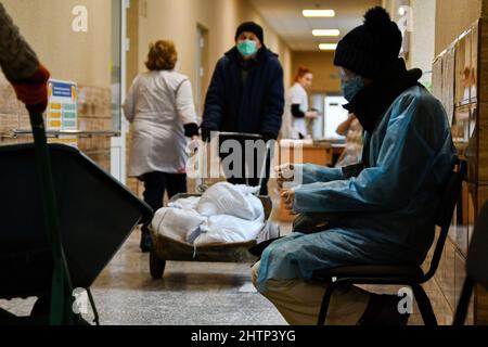 Kramatorsk, Ucraina, 1/3/2022, Un paziente è visto seduto sul banco d'attesa come lavoratori trasportare sacchi di sabbia per coprire la finestra per la protezione nel Kramatorsk ospedale città. Il presidente russo Vladimir Putin ha ordinato l'intervento militare il 24 febbraio, giorni dopo aver riconosciuto due enclavi separatisti nell'Ucraina orientale. Egli sosteneva che Mosca non aveva alcun piano per occupare il paese vicino, ma voleva “smilitarizzare” e “denazificare” l’Ucraina. Il presidente ucraino Volodymyr Zelenskyy ha accusato la Russia di cercare di installare un governo di burattini e ha detto che gli ucraini difenderanno il loro paese contro i Russi Foto Stock