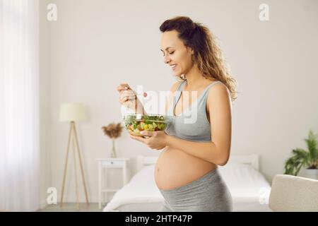 Giovane bella donna incinta mangiare insalata di verdure fresche verdi al mattino in camera da letto. Foto Stock