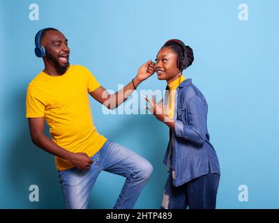 Coppia gioiosa che suona la chitarra aerea e ascolta musica con le cuffie, creando una divertente performance di danza in studio. Uomo e donna innamorati divertirsi con strumenti acustici e sonore sulle cuffie. Foto Stock