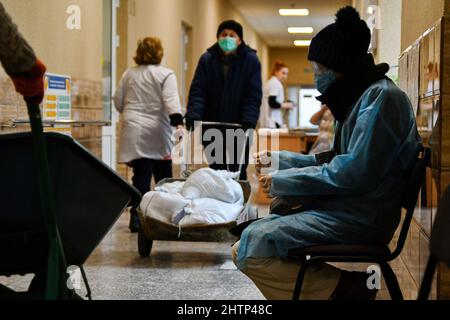 Un paziente è visto seduto sul banco d'attesa mentre i lavoratori trasportano sacchi di sabbia per coprire la finestra per la protezione nell'ospedale della città di Kramatorsk. Il presidente russo Vladimir Putin ha ordinato l'intervento militare il 24 febbraio, giorni dopo aver riconosciuto due enclavi separatisti nell'Ucraina orientale. Egli sosteneva che Mosca non aveva alcun piano per occupare il paese vicino, ma voleva “smilitarizzare” e “denazificare” l’Ucraina. Il presidente ucraino Volodymyr Zelenskyy ha accusato la Russia di cercare di installare un governo di burattini e ha detto che gli ucraini difenderanno il loro paese contro l'aggressione russa. (Foto di Andri Foto Stock