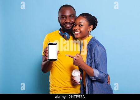 Coppia sorridente che punta a uno schermo bianco vuoto sullo smartphone in studio. Fidanzata e fidanzata che usa il cellulare con modello di chiave chroma e spazio copia mockup. Persone moderne Foto Stock