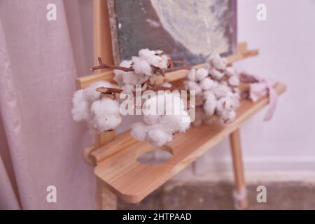 ramo di una pianta di cotone giace su un cavalletto di legno. Design interno dello spazio domestico dell'artista creativo. Foto Stock