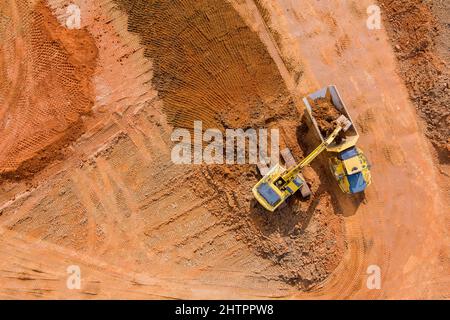 La vista aerea del bulldozer sta raschiando uno strato di terreno e l'escavatore sta scavando e scaricando terra per il carico sui dumper Foto Stock