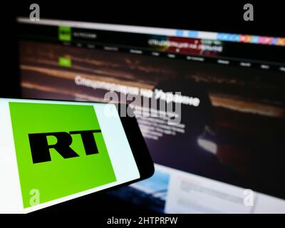 Telefono cellulare con logo della compagnia televisiva russa controllata dallo stato RT sullo schermo di fronte al sito web. Messa a fuoco al centro-sinistra del display del telefono. Foto Stock