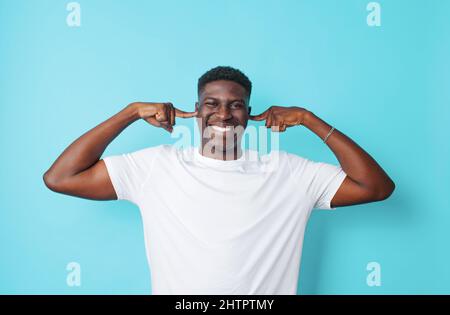 Un bel ragazzo americano in una T-shirt bianca copre le orecchie con le dita su uno sfondo isolato blu. Foto Stock