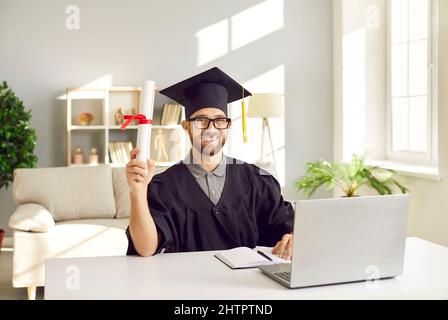 Felice studente che ha ottenuto il suo grado in linea seduto alla scrivania, tenendo diploma e sorridente Foto Stock