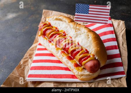 Primo piano della bandiera americana su hot dog con salsa di pomodoro e mostered su carta di cera marrone a tavola Foto Stock