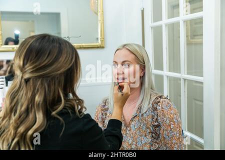 Cosmetologist che applica il trucco del rivestimento del labbro ad una donna bionda in una sedia di trucco Foto Stock