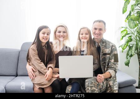 Un uomo militare felice sorridendo e abbracciando la sua famiglia mentre usa un computer portatile al chiuso. Foto Stock