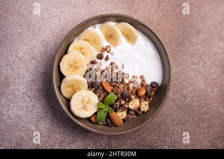 Vista dall'alto della granola con yogurt, banana fresca, gocce di cioccolato e mandorle per una colazione sana su sfondo di cemento marrone Foto Stock