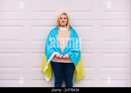 Donna Ucraina matura con capelli biondi gesto serio con bandiera Ucraina blu e gialla su sfondo bianco per strada. Guerra Ucraina, invasione, p. Foto Stock