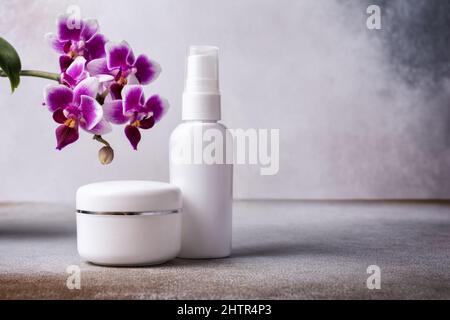 Mockup di prodotti cosmetici per la cura della pelle. Bottiglia bianca di lozione ecologica e vasetto di crema con splendidi fiori di orchidea viola su sfondo grigio moderno Foto Stock