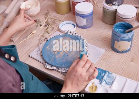 Vasaio professionale lavora su piastre di pittura in officina. Donna ceramista dipinge un piatto di colore blu. Foto Stock