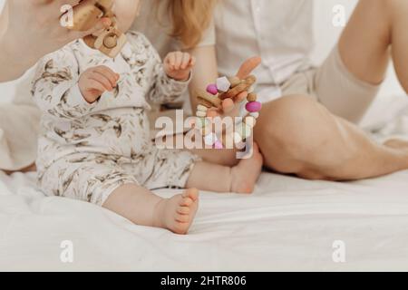Vista croppata della famiglia con pezzetto bambino bambino piccolo che tiene moderno legno ecologico massaggiante seduta su letto bianco. Foto Stock