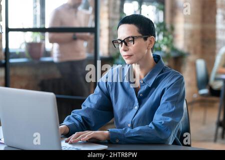 Donna d'affari seria con i capelli corti negli occhiali che battono sul laptop mentre si siede al suo posto di lavoro in ufficio Foto Stock
