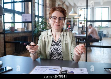 Ritratto di giovane donna d'affari in occhiali guardando la macchina fotografica mentre si siede al suo posto di lavoro e lavorando con grafici finanziari Foto Stock