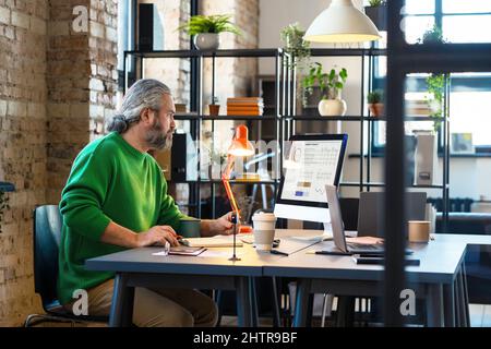 Uomo d'affari maturo bearded che guarda il monitor del calcolatore con i grafici mentre si siede al suo posto di lavoro in ufficio, lui che analizza le statistiche Foto Stock