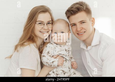 Ritratto di giovane felice sorridente bella famiglia con cherubico occhio blu bambino bambino toddler in vestiti bianchi seduti. Foto Stock