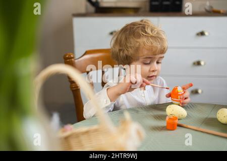 Ragazzino con capelli ricci biondi dipinge le uova di Pasqua con un pennello Foto Stock