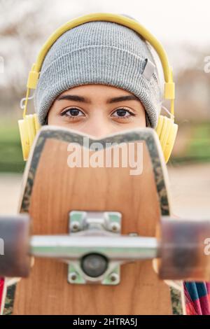 Ritratto del giovane sportivo ispanico con cappuccio e cuffie guarda la fotocamera e copre il suo volto con skateboard. Foto Stock