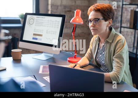 Donna d'affari seria in occhiali seduti al tavolo con il computer durante la presentazione alla riunione Foto Stock